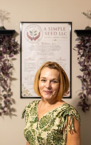 Cassandra Myers smiling in front of the framed welcome sheet in her treatment room, trailing plants on shelves either side